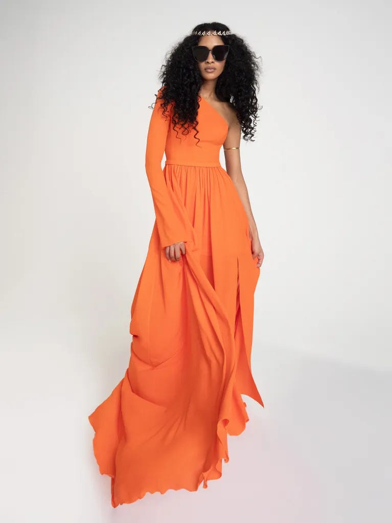 Woman with curly hair, wearing an orange long evasee party dress with asymmetrical neckline, against a grey background.